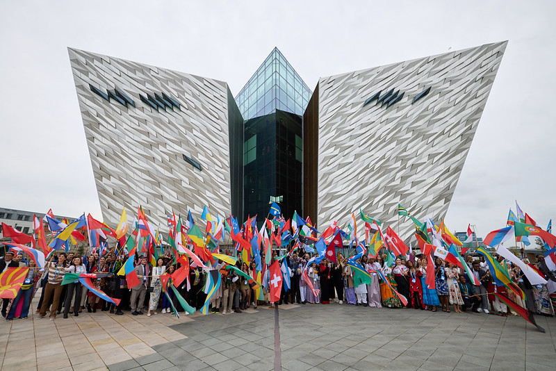 Group photo of One Young World attendees holding national flags in front of the Titanic Belfast building