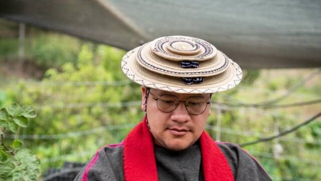 Leider Andres Tombe Morales wearing round, ornate hat, red neck garment amongst waist-to-shoulder-high shrubs