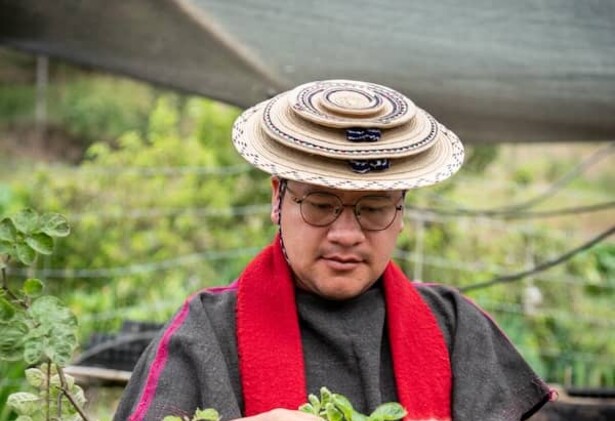 Leider Andres Tombe Morales wearing round, ornate hat, red neck garment amongst waist-to-shoulder-high shrubs